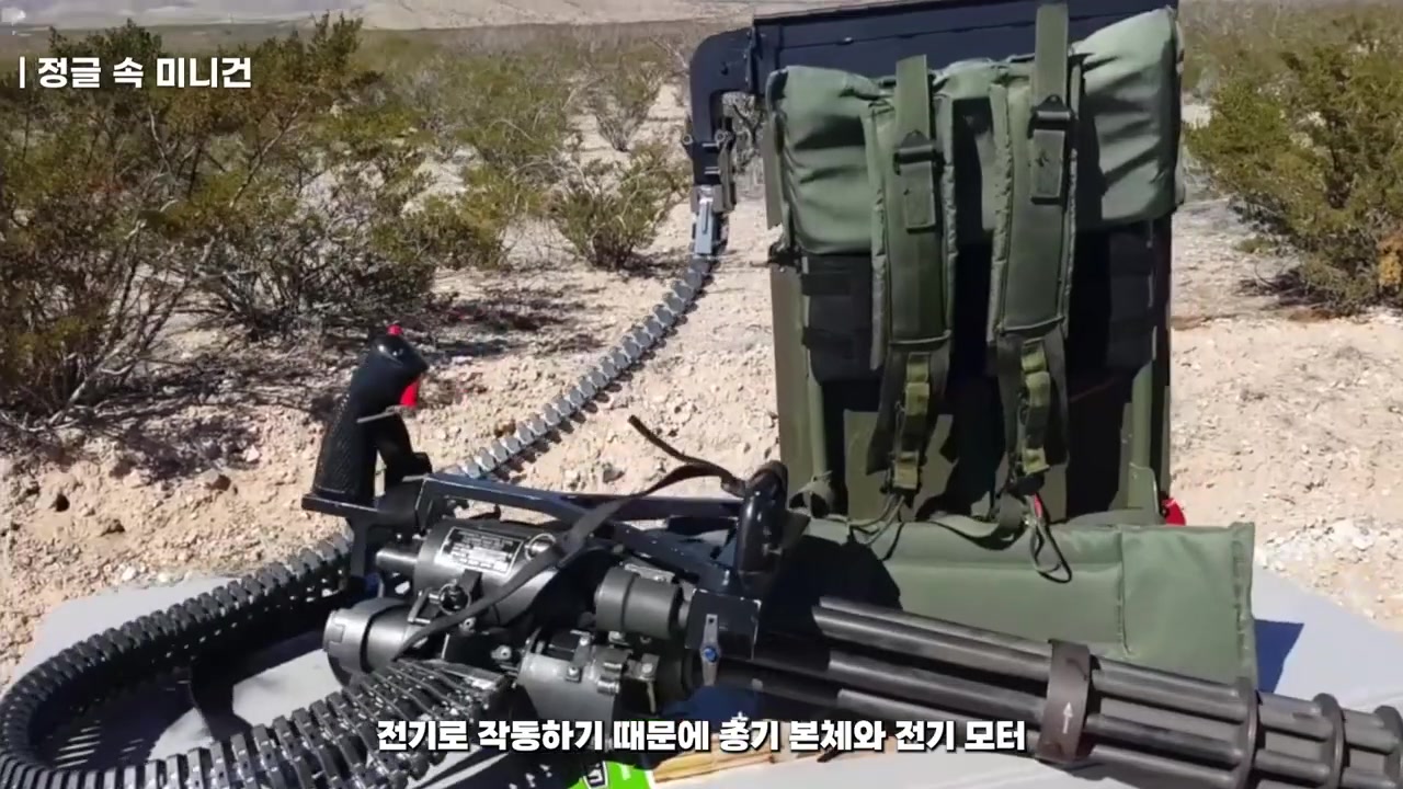 사막에서 삼각대 없이 놓인 휴대용 미니건, 초록색 전기모터와 대형 배터리팩이 스트랩으로 고정된 모습