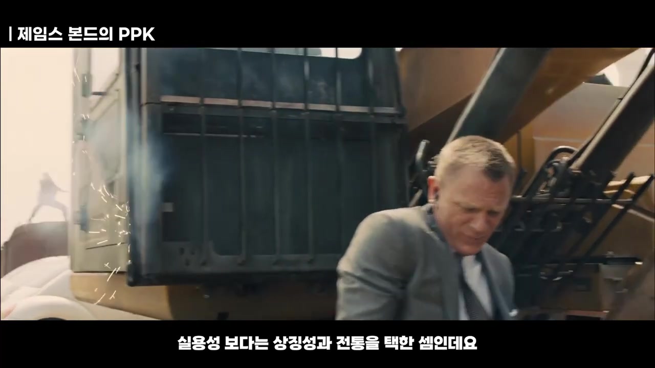 정장을 입은 인물이 화물 컨테이너 옆에서 불꽃이 튀는 장면 앞에 서 있는 모습